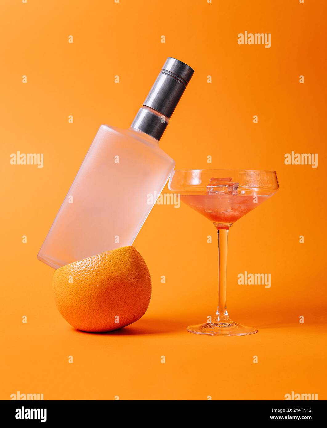 Wodka-Flasche, die auf einer Orange neben einem Cocktailglas mit Eiswürfeln und einem rosafarbenen Cocktail auf einem orangen Hintergrund balanciert Stockfoto