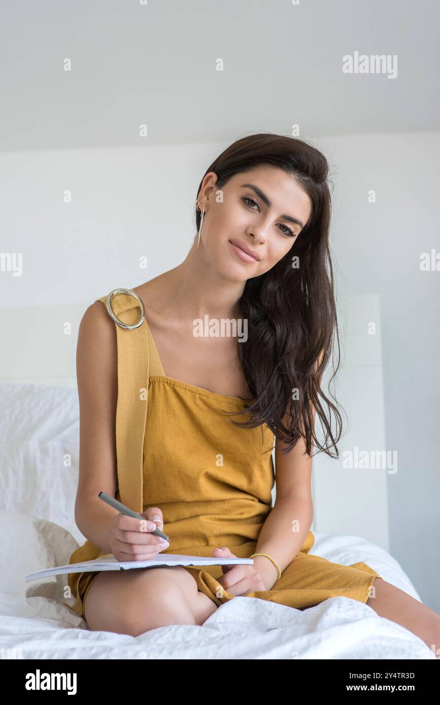Junge Frau macht Notizen im Bett. Sie ist ein braunes Kleid und hat lange schwarze Haare. Stockfoto