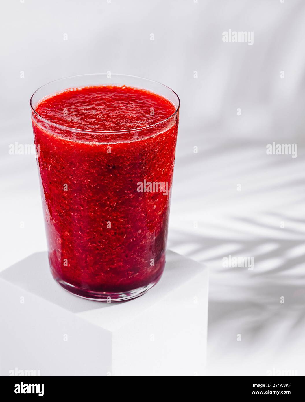 Ein Glas roter Beeren-Smoothie sitzt auf einem weißen Würfel mit tropischem Pflanzenschatten Stockfoto