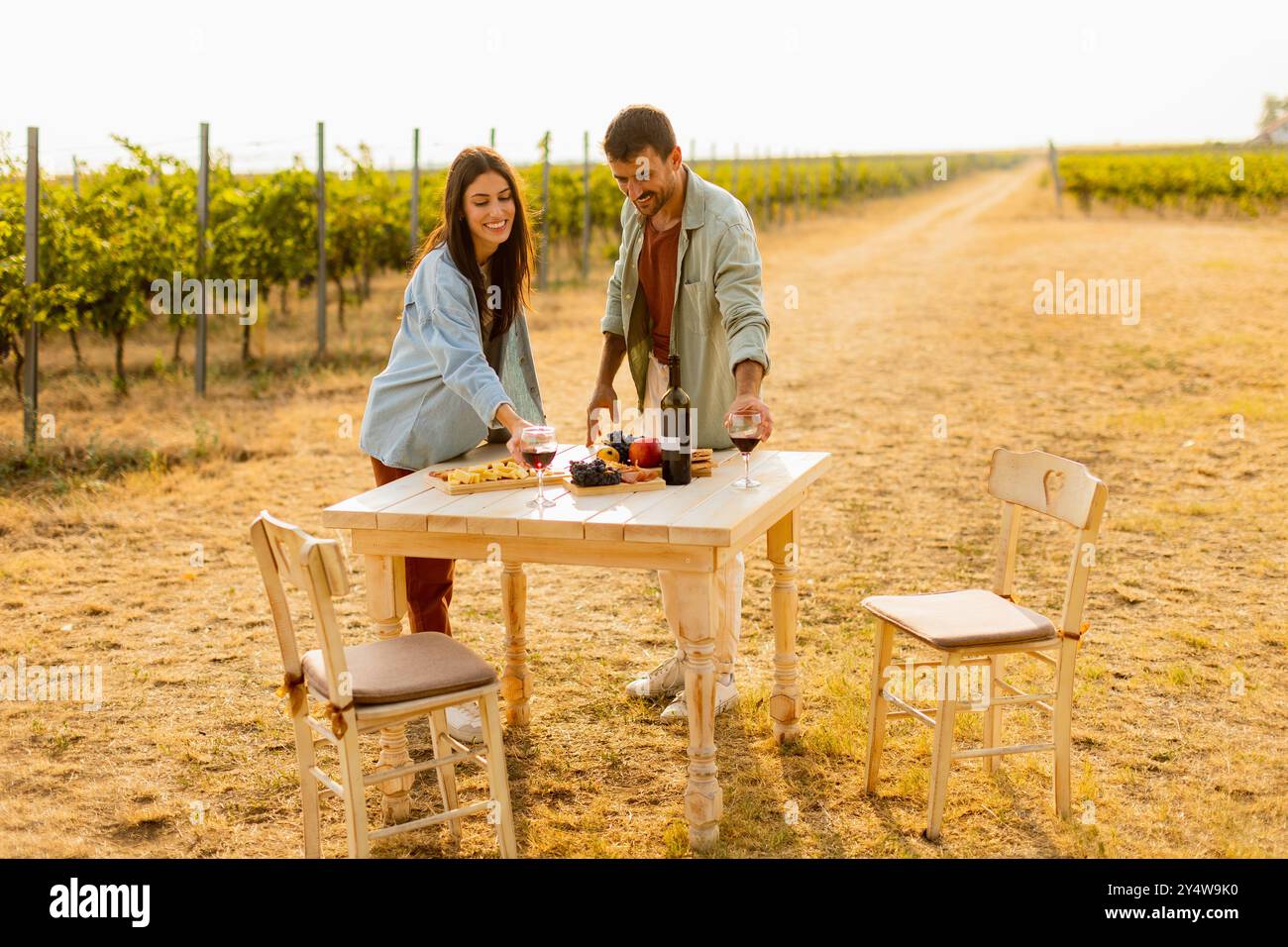 Im warmen Sonnenlicht begrüßt ein fröhliches Paar ein köstliches Picknick zwischen den Weinreben und genießt Wein und Gourmet-Leckereien Stockfoto