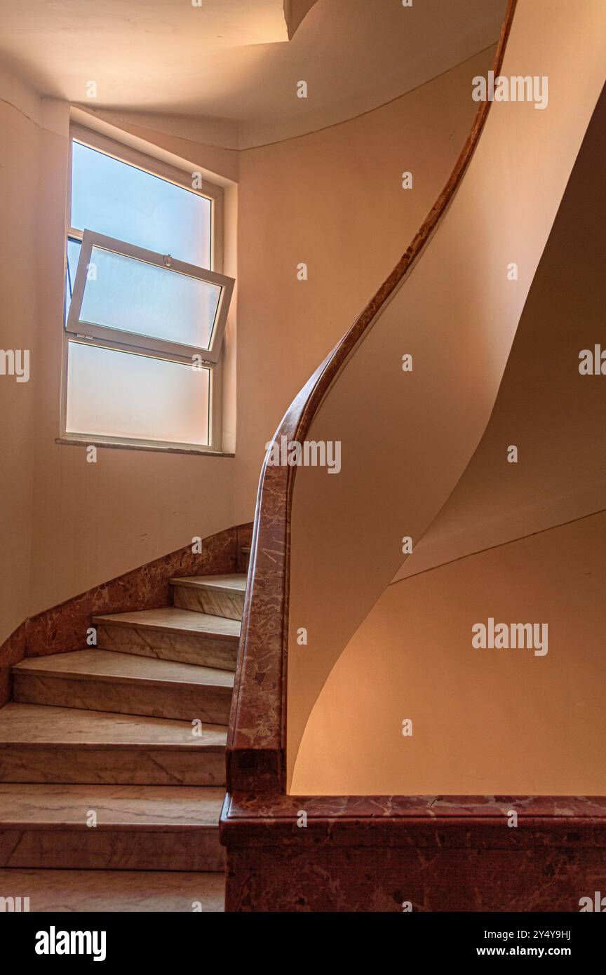 Eine gewundene Wendeltreppe mit Marmortreppen und bannister, mit pfirsichfarbenen Wänden und einem zerrissenen Fenster mit einem blauen Himmel. Stockfoto