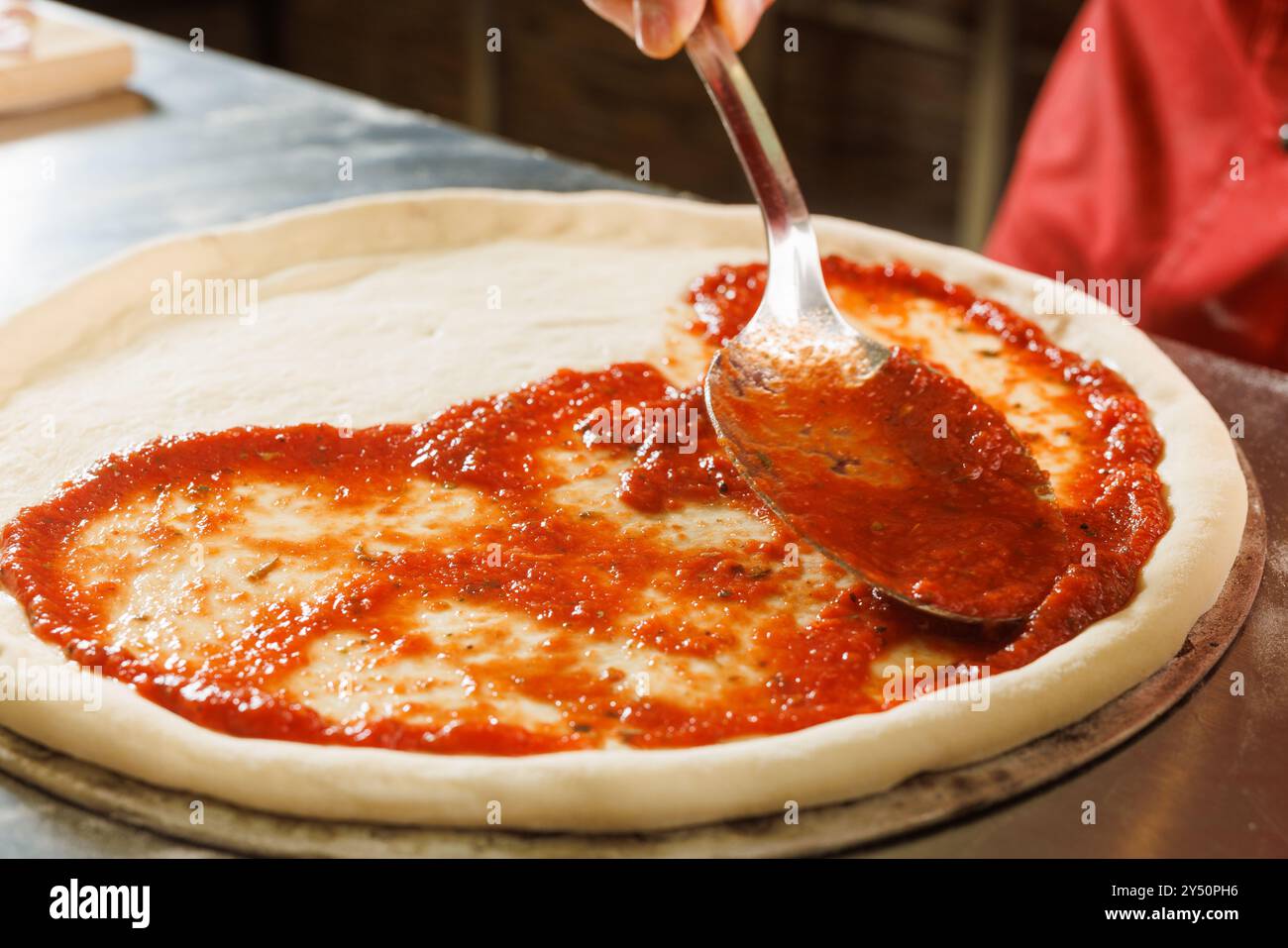 Eine Person, die Tomatensauce auf Pizzateig mit einem Kelle in einer Küche verteilt. Stockfoto