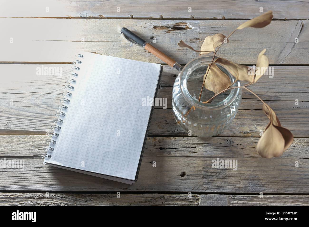 Leeres Spiral Notizblock Mockup, Stift und eine Glasvase mit trockenen braunen Blättern auf einem rustikalen Tisch aus verwitterten Holzdielen, Herbst- und Winterstill liv Stockfoto