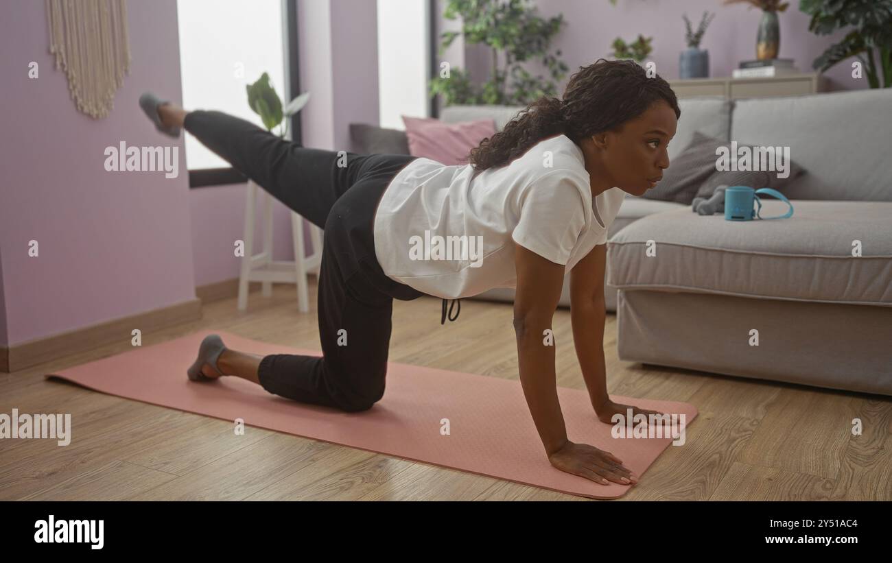 Eine junge afroamerikanische Frau übt Yoga auf einer Matte in ihrem gemütlichen Wohnzimmer und zeigt einen konzentrierten und entschlossenen Ausdruck beim Training Stockfoto