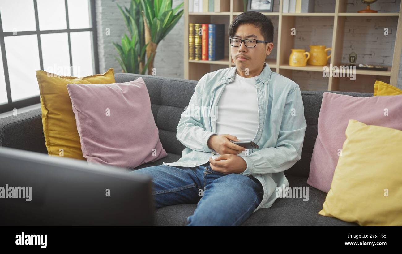 Ein junger asiatischer Mann sitzt lässig auf einer Couch, entfernt in der Hand, in einem gemütlichen Wohnzimmer. Stockfoto