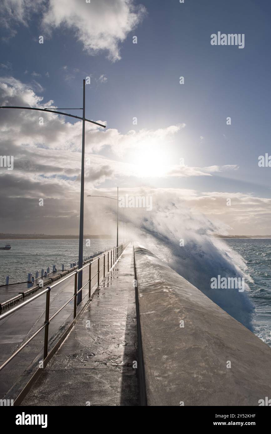 Große Wellen krachen über den Pier, Sonnenlicht bricht durch Wolken Stockfoto