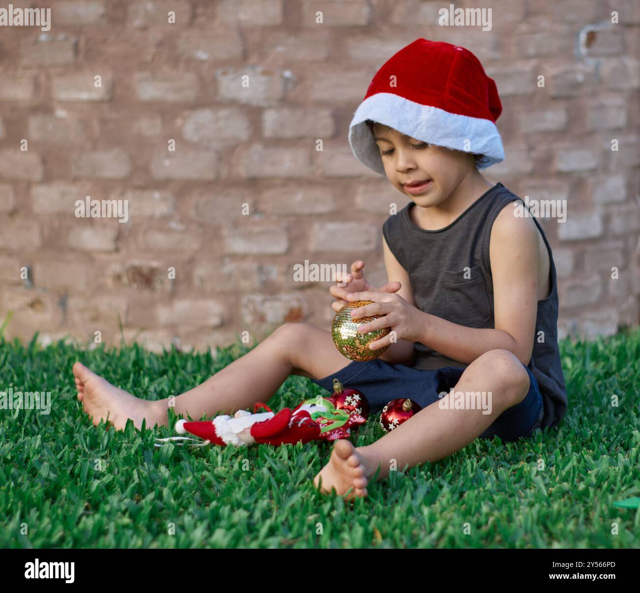 latino Boy sitzt auf dem grünen Rasen und spielt mit weihnachtsdekorationen, trägt ein Sommerkleid mit einem Muskelhemd und Shorts und trägt einen roten sa Stockfoto