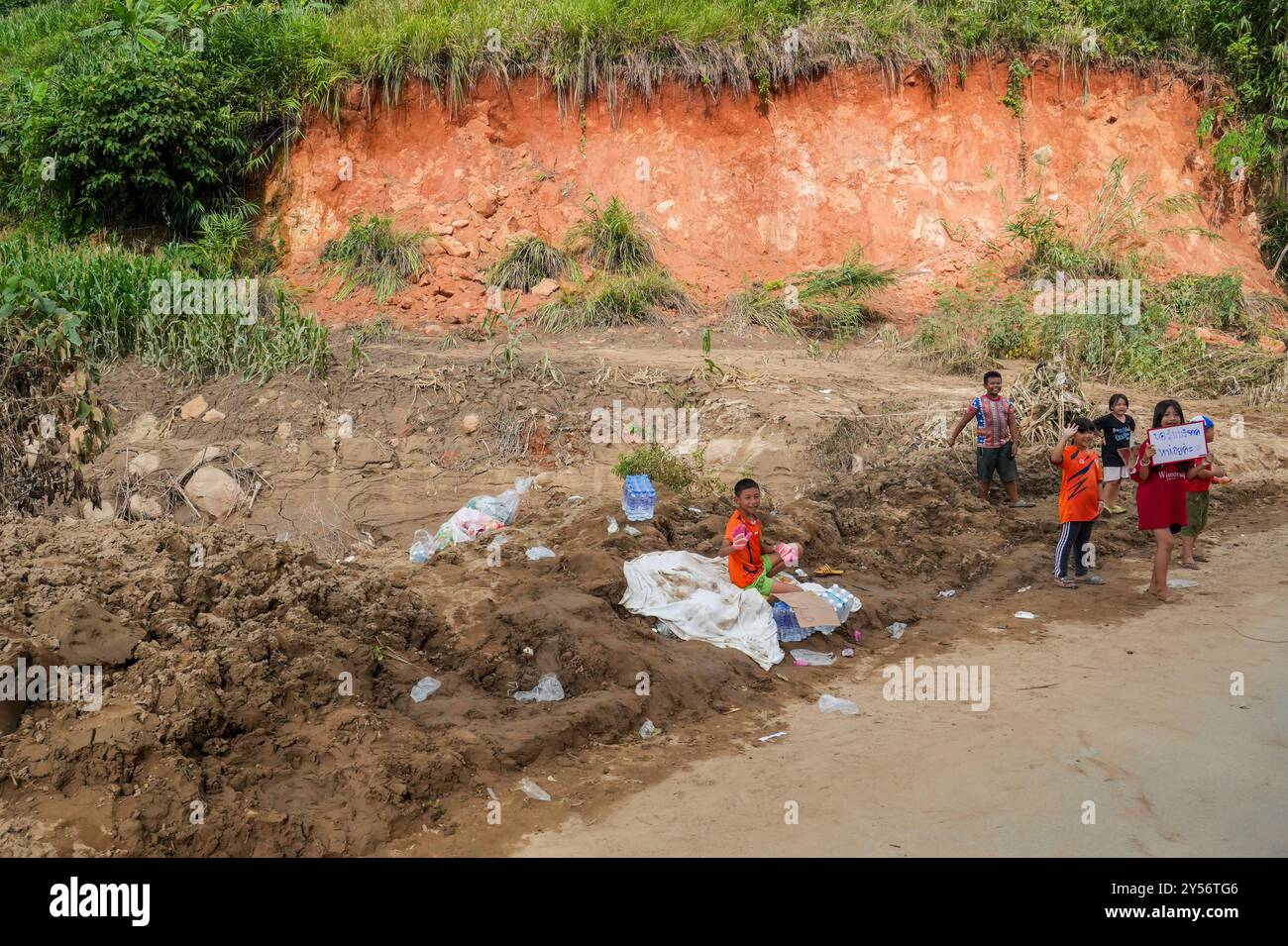 Erwachsene und Kinder werden auf einer Straße in den Bergen, im Unterbezirk Mae Yao, nach der Überschwemmung in Chiang Rai, Thailand, mit einem Plakat gesehen, das um Hilfe und Spenden bittet. Nach den schlimmsten Überschwemmungen seit 80 Jahren werden in Chiang Rai Restaurierungsbemühungen unternommen, wobei die Menschen die umfassende Säuberung von dickem Schlamm und Schutt in Angriff nehmen, der das Gebiet überholt hat. Viele Bewohner wurden aufgrund strukturell unsicherer Häuser zur Evakuierung gezwungen, was eine Spur der Zerstörung durch die Überschwemmungen hinterließ. Stockfoto