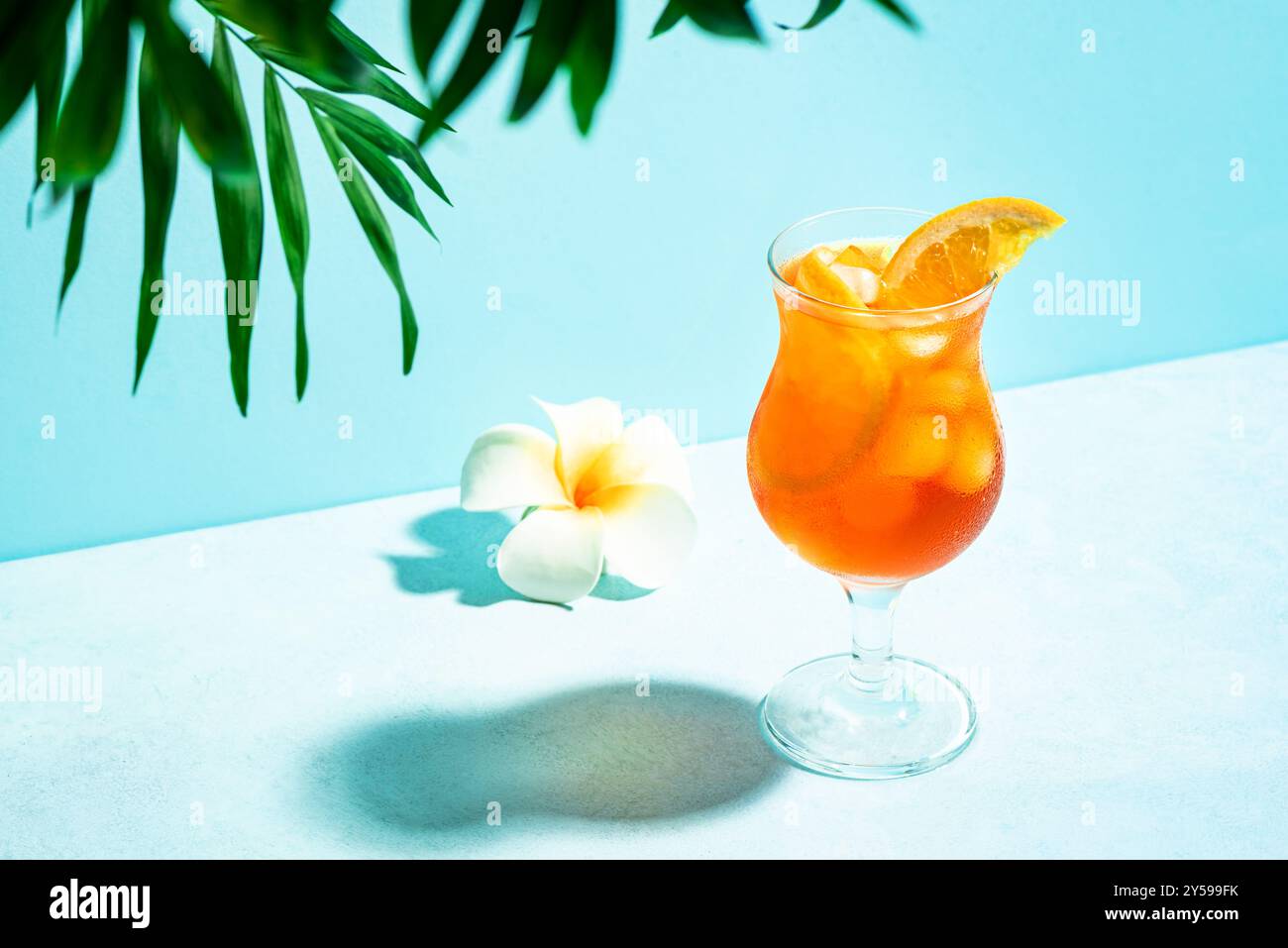 Roter alkoholischer Sommercocktail mit Orangenscheiben, Blume und Palmblatt auf blauem Hintergrund, Kopierraum. Trendiger Mai Tai Cocktail, Strand entspannen und Stockfoto
