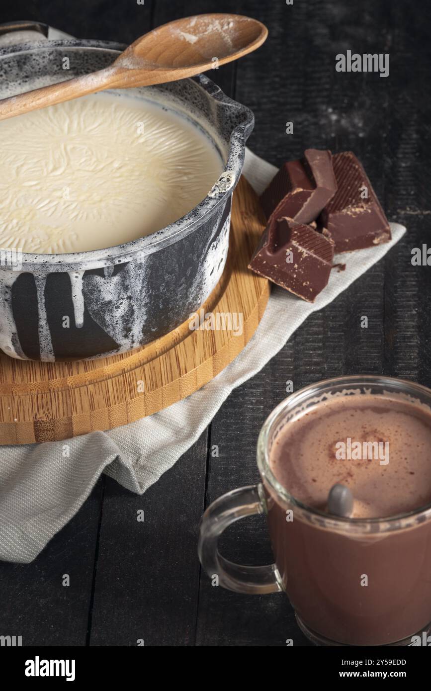 Milch in einem schwarzen Topf gekocht, über die Kanten verteilt, Schokoriegel und ein Glasbecher mit heißer Schokolade auf einem rustikalen Tisch Stockfoto