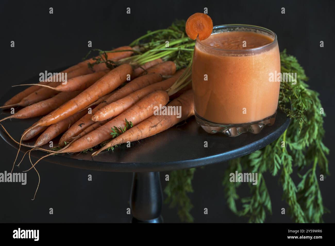 Vegetarisches und diätetisches Konzept mit einem Glas frischen Karottensaft und einem Haufen frisch geernteter Karotten, auf einem schwarzen runden Tisch, bei schlechtem Licht Stockfoto