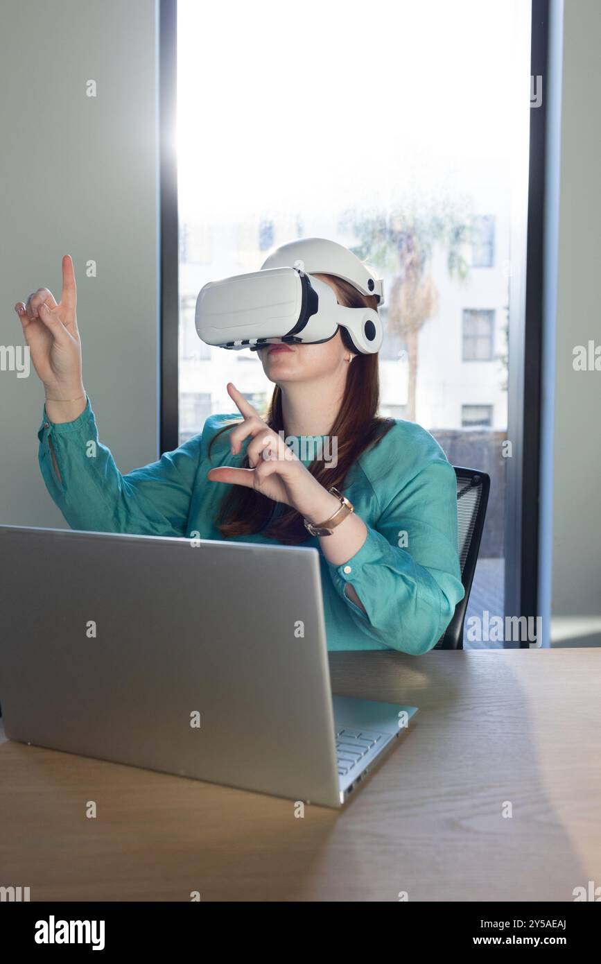 Im Büro mit VR-Headset, Frau, die mit der virtuellen Umgebung interagiert, während sie an einem Laptop arbeitet Stockfoto