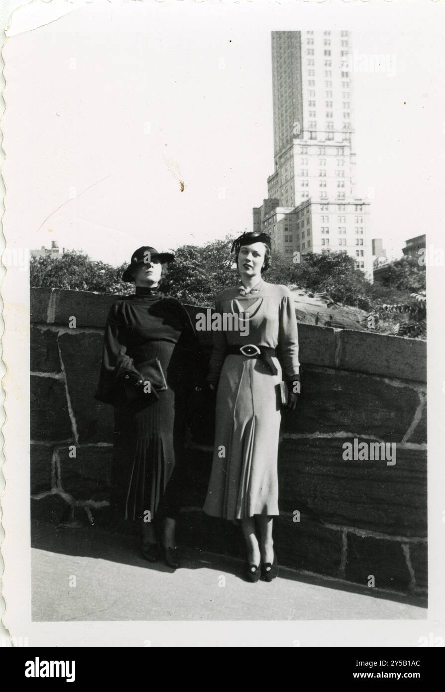 Zwei elegant gekleidete Frauen posieren vor einer Skyline der Stadt und zeigen Mode aus den 1930er Jahren mit strukturierten Kleidern, Gürtelbündchen und stilvollen Hüten. Das Foto fängt die Raffinesse und urbane Eleganz der Epoche ein Stockfoto