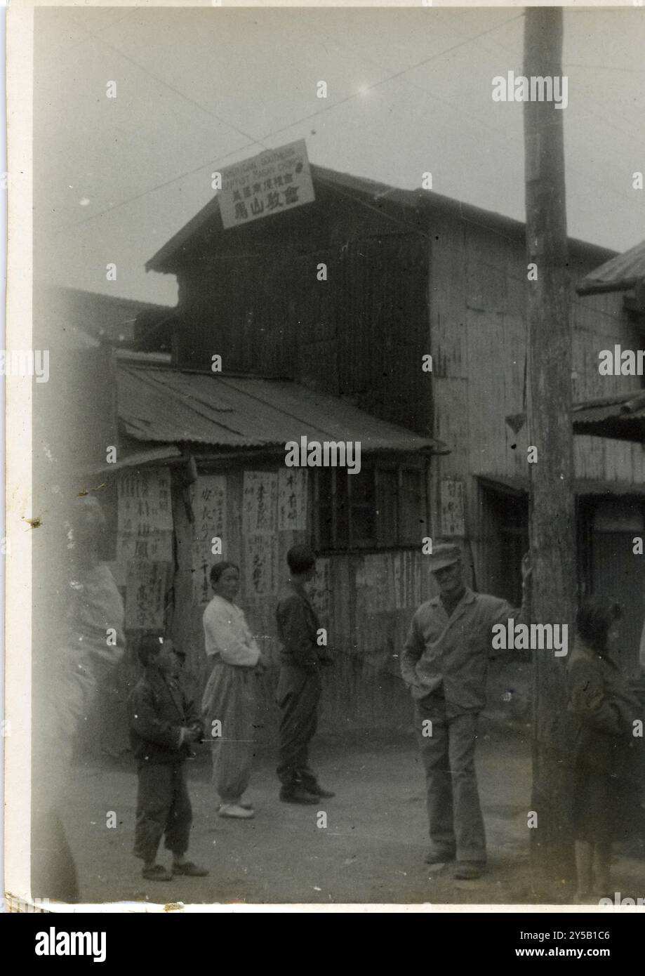 Amerikanischer GI posiert für ein Foto auf einem geschäftigen Markt mit Straßenverkäufern und Fußgängern in Japan nach dem Zweiten Weltkrieg. Im Hintergrund befindet sich eine Baptistenkirche. Stockfoto