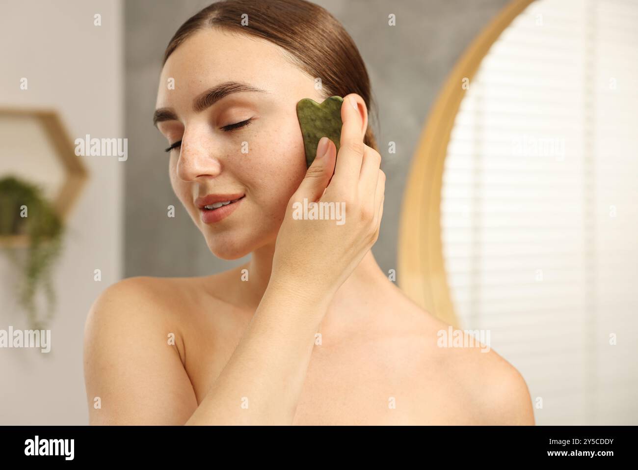 Schöne junge Frau, die eine Gesichtsmassage mit Gua-Sha-Werkzeug drinnen macht Stockfoto