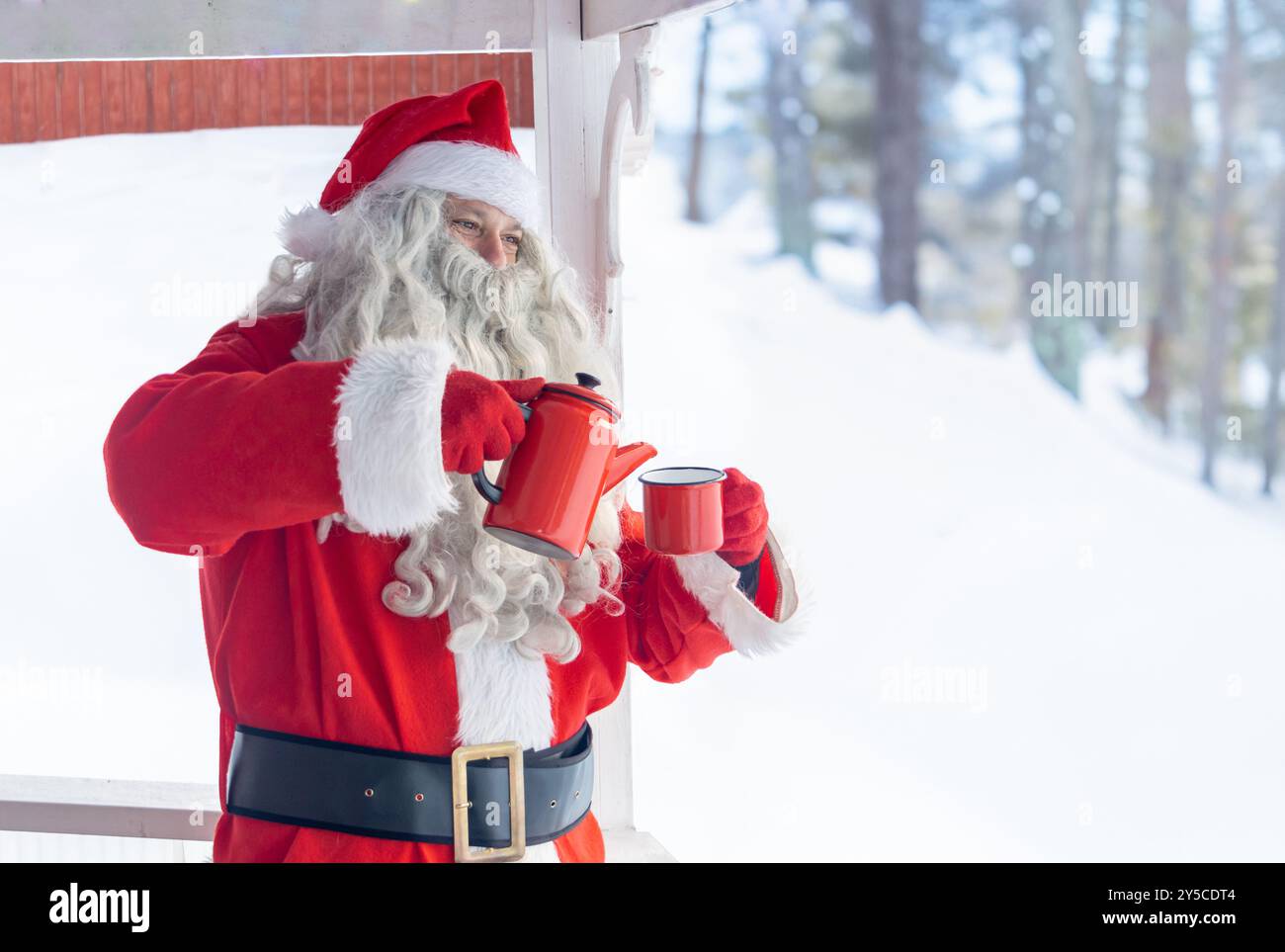 Der Weihnachtsmann schenkt sich ein heißes Getränk auf der Terrasse eines Hauses in der Nähe eines verschneiten Waldes Stockfoto