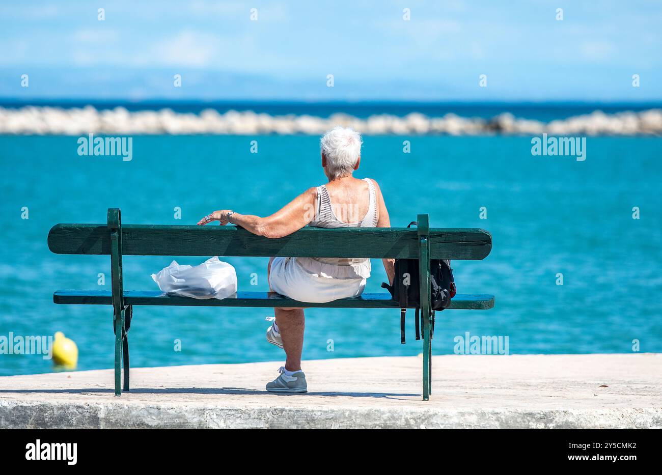 Ältere reife Frau, Dame, die auf einer Bank sitzt und tief in Gedanken aufs Meer blickt, Dame, die allein auf der Bank sitzt und tagträumt, reife Frau allein auf der Bank. Stockfoto