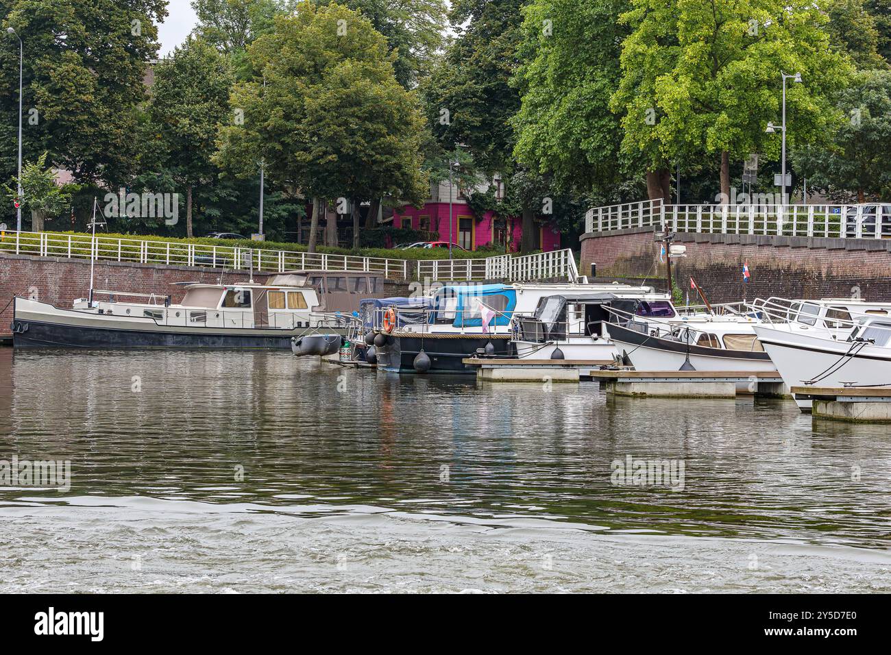 Historischer alter Binnenhafen von Maastricht mit Schiffen, die am Kai ankern, Rampe mit Metallgeländer und üppigen Laubbäumen im Hintergrund, bewölkter Tag i Stockfoto