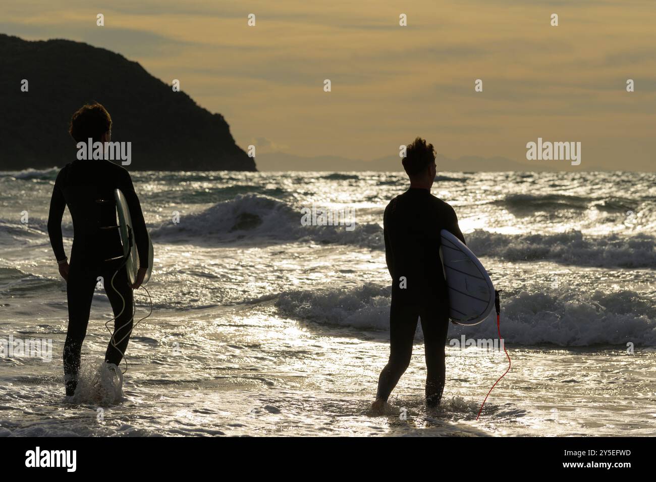 Zwei junge Männer mit Surfbrettern bekämpfen die Wellen bei Sonnenuntergang im Golf von Baratti, Piombino, Livorno, Italien Stockfoto