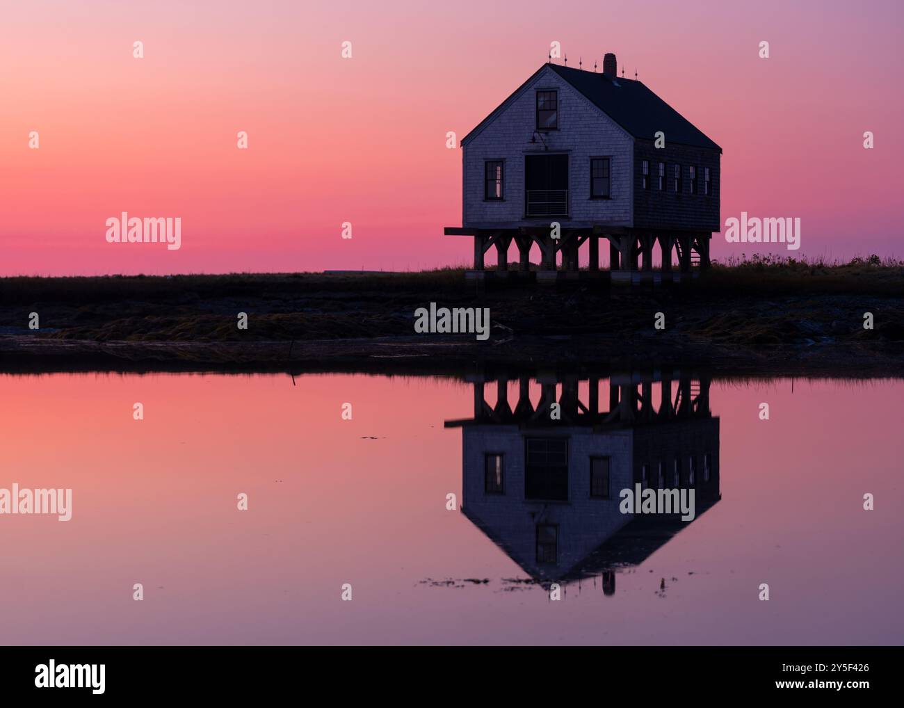Ein auf Stelzen stehendes Haus spiegelt sich perfekt in dem ruhigen Wasser darunter wider. Der Himmel ist mit einem Farbverlauf aus rosa und violetten Tönen bemalt, was auf eine Ethik hinweist Stockfoto