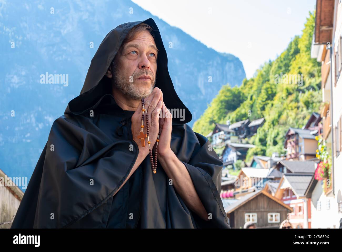 Ein älterer bärtiger Mönch in einer schwarzen Kapuze hält einen Gebetsrosar, während er vor einem verschwommenen Hintergrund aus Bergen und einem Bergdörfer steht Stockfoto