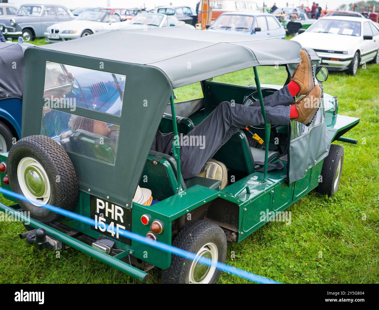 MINI MOKE MIT EINEM MANN, DER ENTSPANNT EIN BUCH AUF EINEM TABLET MIT STIEFELN AUF EINEM KLASSISCHEN SAINT MAWGAN DAMPFRALLYE LIEST Stockfoto