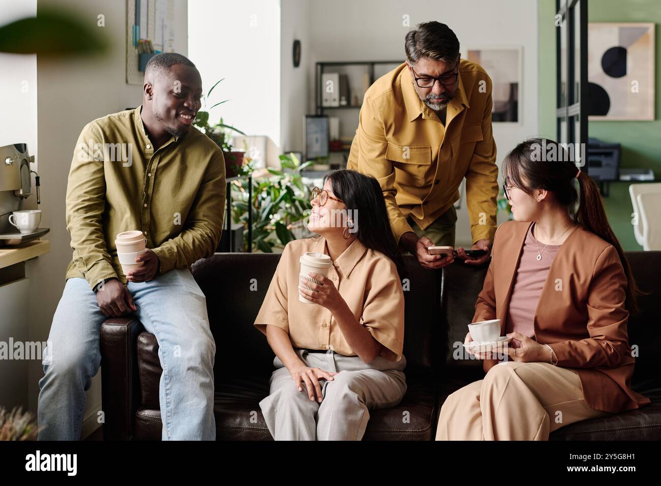 Gruppe verschiedener Kollegen, die gerne Gespräche führen, während sie Kaffeetassen in einem modernen Büro halten. Entspannte Atmosphäre mit komfortablen Sitzgelegenheiten und viel Grün, die eine ungezwungene Interaktion fördert Stockfoto