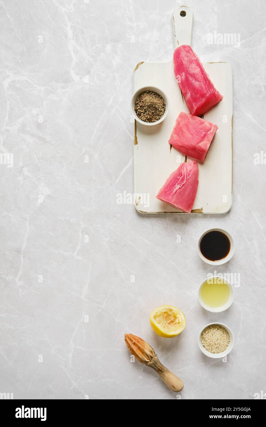 Blick von oben auf frisch geschnittene Thunfischfilets, die auf einem weißen Schneidebrett angeordnet sind, zusammen mit verschiedenen Gewürzen und einer gepressten Zitrone, bereit für die Zubereitung i Stockfoto
