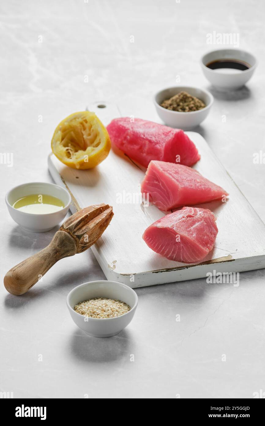 Frische Thunfischsteaks werden auf einem Schneidebrett zusammen mit gepresster Zitrone, Olivenöl und kleinen Schüsseln mit verschiedenen Gewürzen zubereitet, die in A k zubereitet werden können Stockfoto