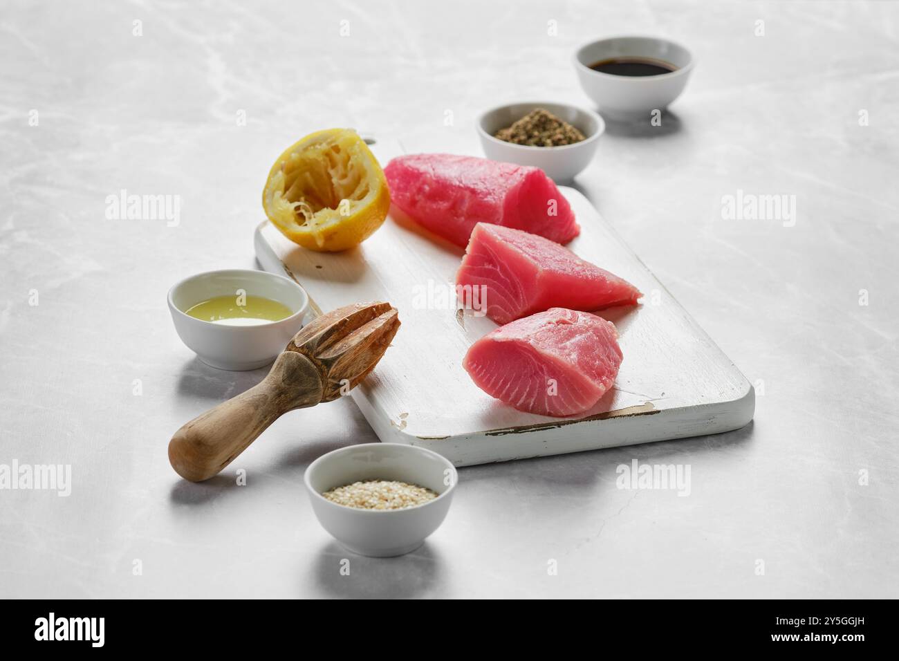 Auf der hell beleuchteten Arbeitsplatte finden Sie frische Thunfischfilets, eine gepresste Zitrone und verschiedene Gewürze, die für ein köstliches kulinarisches Erlebnis bereit sind Stockfoto
