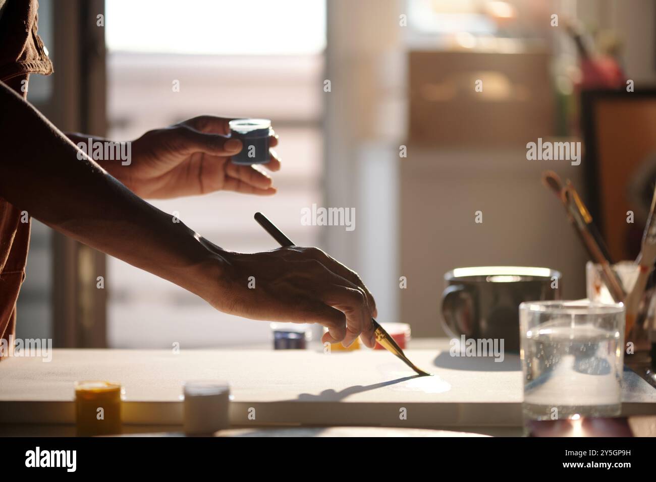 Handpinsel in hellem Studio mit verschiedenen Kunstgegenständen auf dem Schreibtisch. Sonnenlicht, das durch das Fenster strömt, schafft eine warme Atmosphäre in kreativer Umgebung Stockfoto