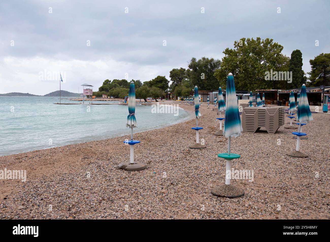 Vodice, Kroatien - 9. September 2024: Gefaltete Sonnenschirme am Blauen Strand an einem bewölkten, windigen Tag ohne Menschen Stockfoto