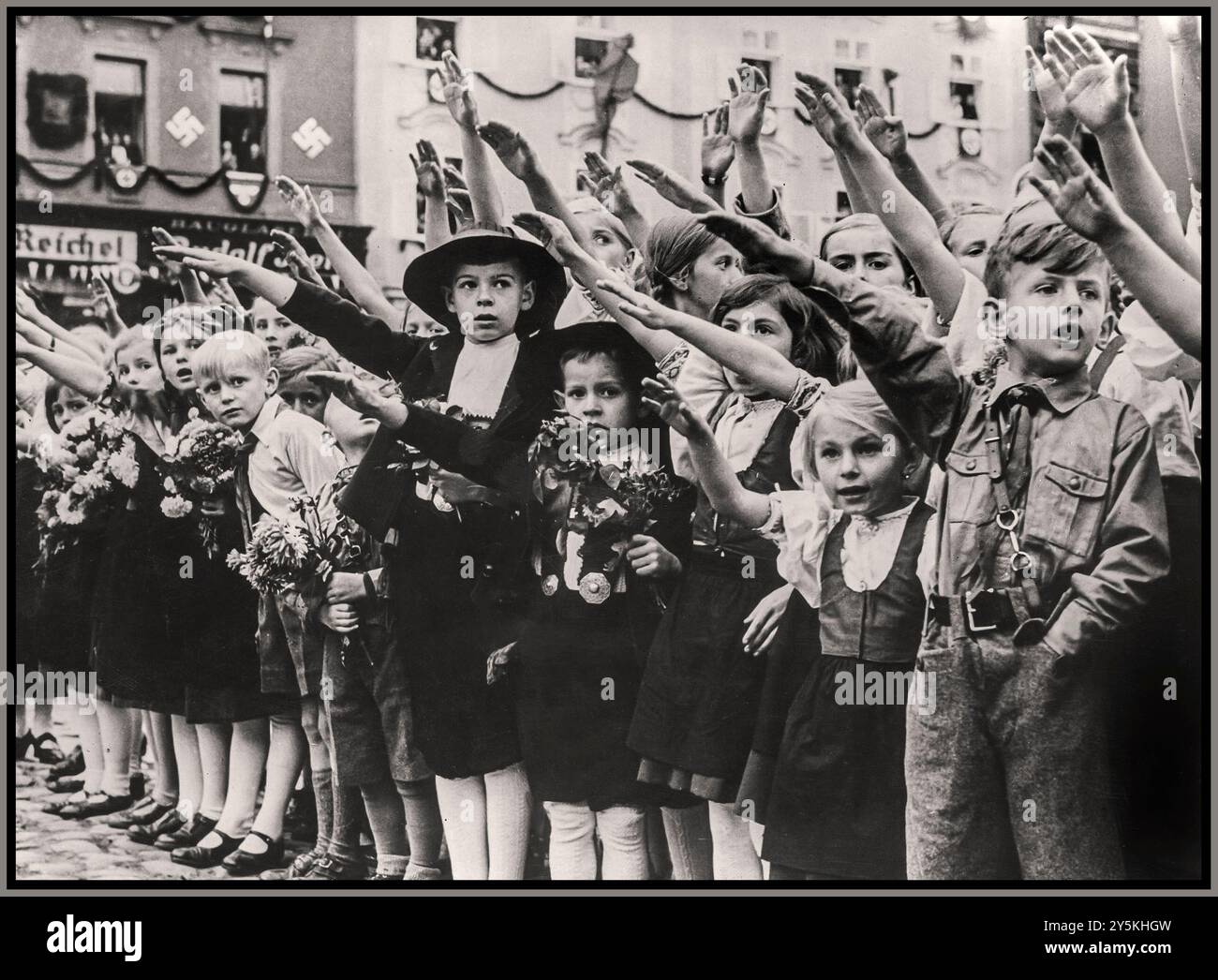 ANSCHLUSS AUSTRIAN CHILDREN HEIL HITLER SALUTE 1930er NS-Propagandabild österreichischer Kinder, darunter ein Hitler-Jugendjunge in Uniform, der Adolf Hitler mit traditionellem Heil Hitler-Salut mit erhobenen Armen begrüßte. Hakenkreuz-Symbol an der Wand im Hintergrund. Wien (Hinweis: Jüdisches Eigentum und später „erworben“ von den Nazis, REICHEL Textilgeschäft in b/g) Wien Österreich Stockfoto