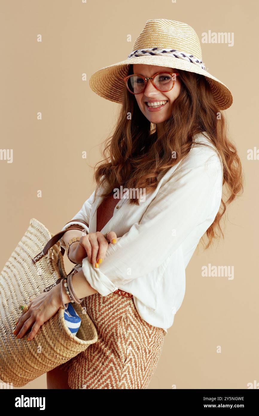 Strandurlaub. Lächelnde elegante Frau mittleren Alters in weißer Bluse und Shorts isoliert auf beigem Hintergrund in Brille mit Strohtasche und Sommerhut. Stockfoto Strandurlaub. Lächelnde elegante Frau mittleren Alters in weißer Bluse und Shorts isoliert auf beigem Hintergrund in Brille mit Strohtasche und Sommerhut. Stockfoto
