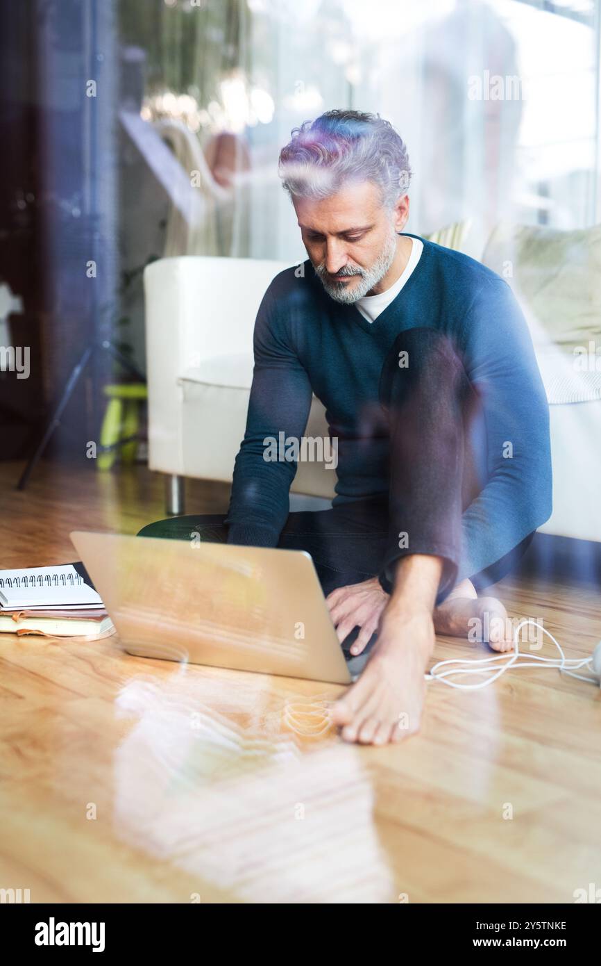 Homeoffice für einen schönen Mann. Mann, der auf dem Boden sitzt und am Laptop arbeitet. Durchgangsfenster. Telearbeit. Stockfoto