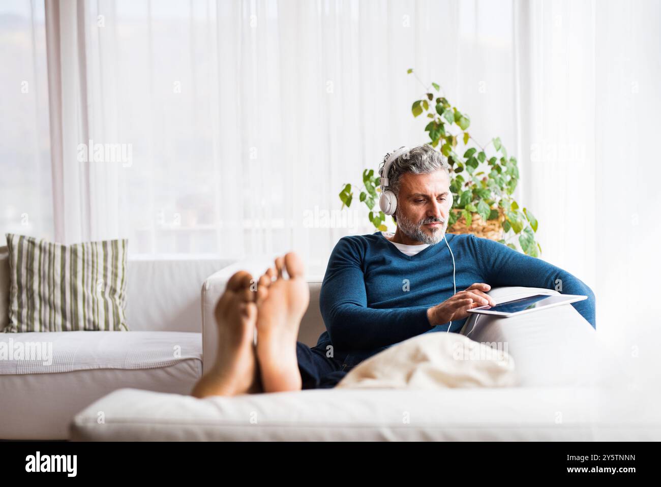 Mann zu Hause, der auf dem Sofa sitzt und ein Tablet hält, Kopfhörer auf dem Kopf hält, Musik hört. Stockfoto