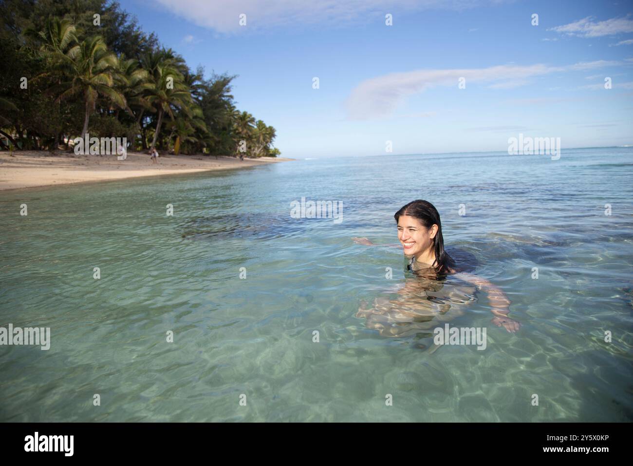 Frau lächelt beim Waten in klarem tropischem Wasser mit Palmen am Strand im Hintergrund, Titikaveka, Rarotonga, Cook Islands Stockfoto