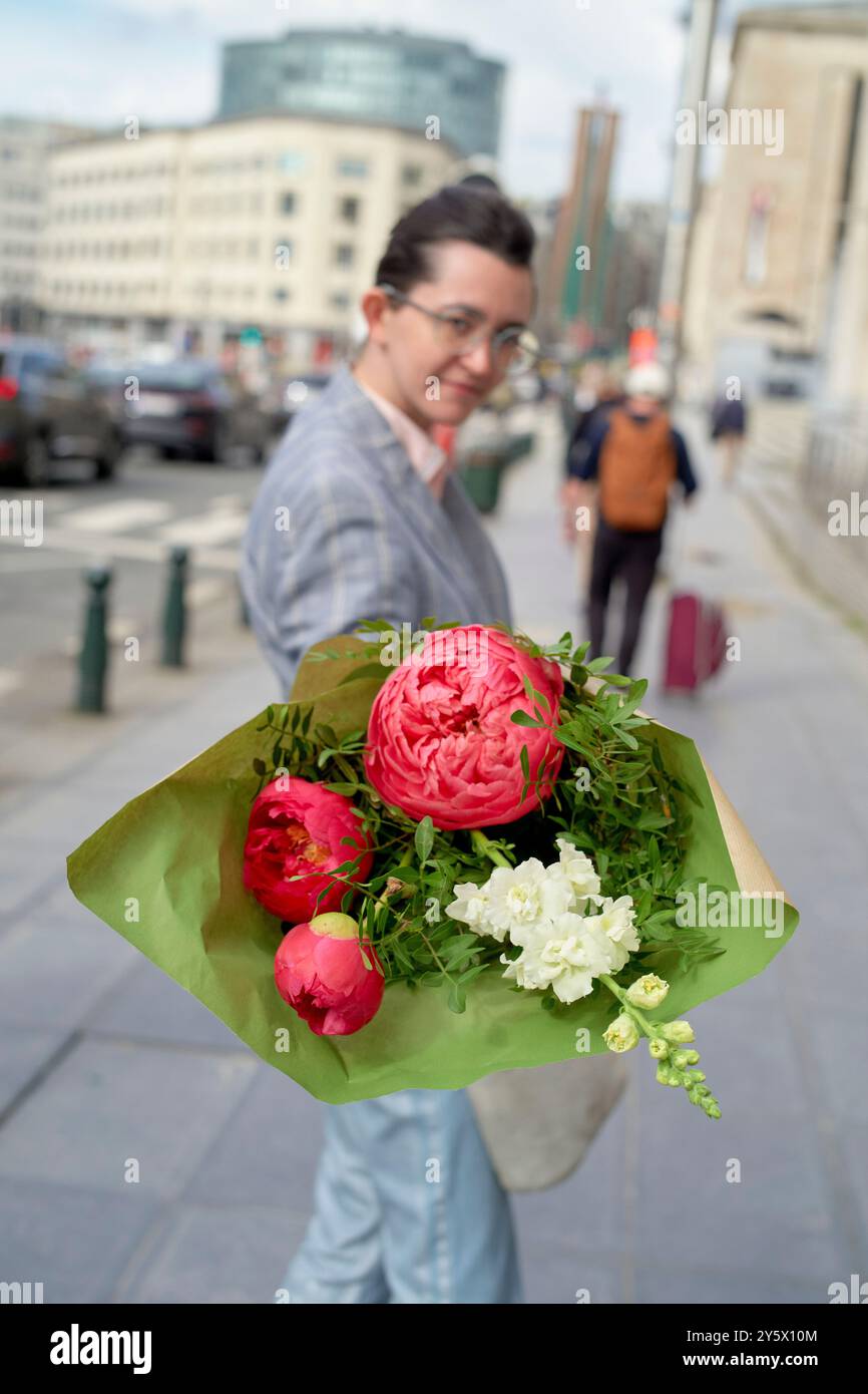 Lächelnde Frau mit Brille, die einen Strauß rosa und weißer Blumen auf einer Stadtstraße hält. Stockfoto