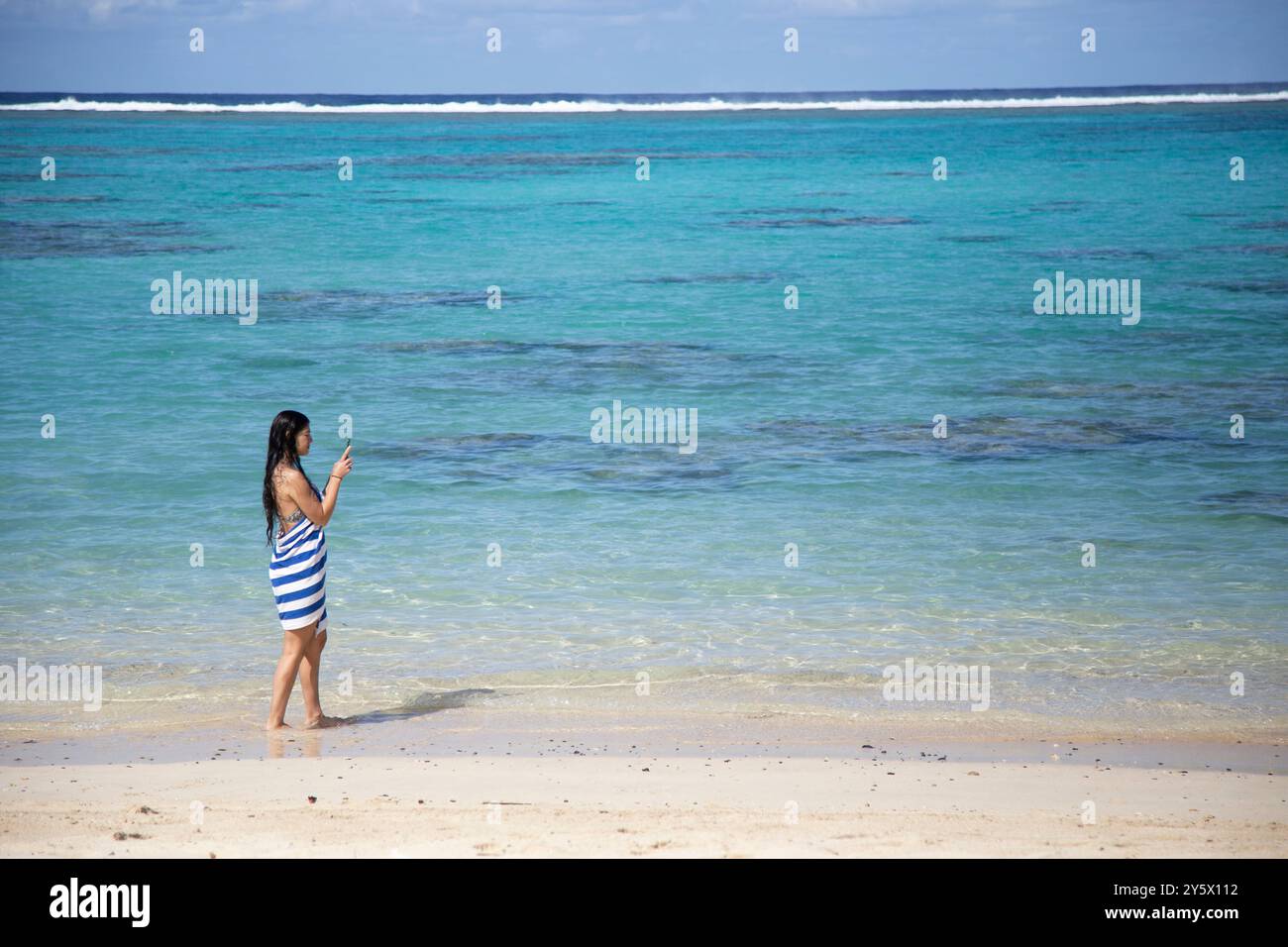 Eine Frau in gestreiftem Badeanzug steht am Rand eines klaren türkisfarbenen Ozeans und blickt in Richtung Horizont, Titikaveka, Rarotonga, Cook Islands Stockfoto