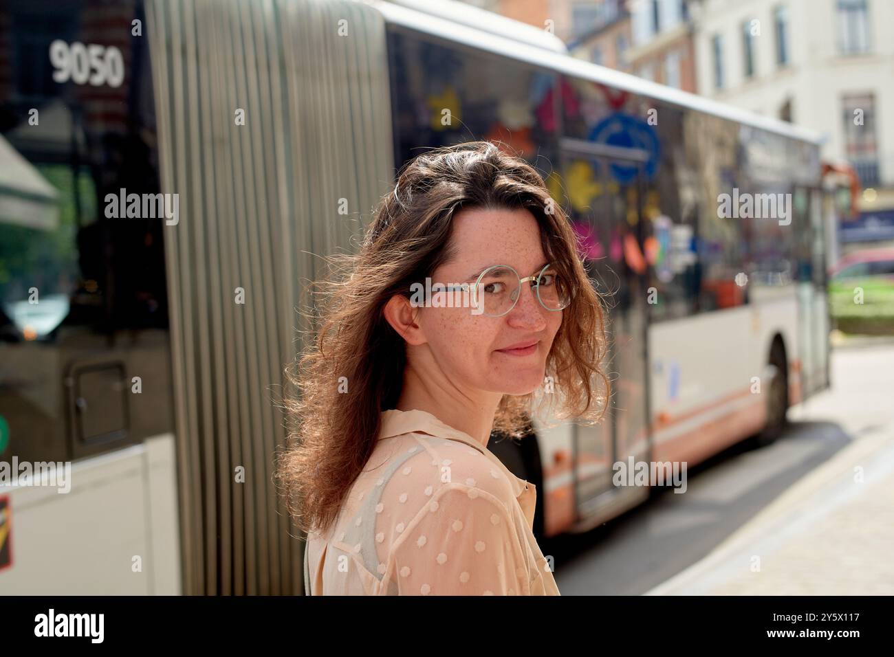 Lächelnde Frau mit Brille, die vor einem fahrenden Bus auf einer sonnigen Stadtstraße steht. Stockfoto