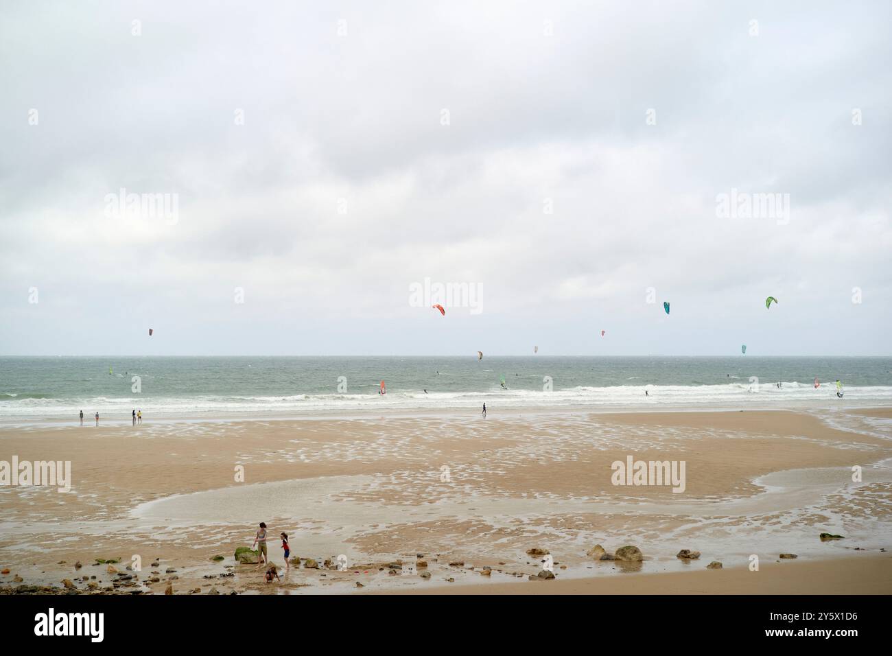 Bewölkter Tag am Strand mit Individuen, die Kiteboarden und auf dem Sand in der Nähe des Wassers spazieren. Stockfoto