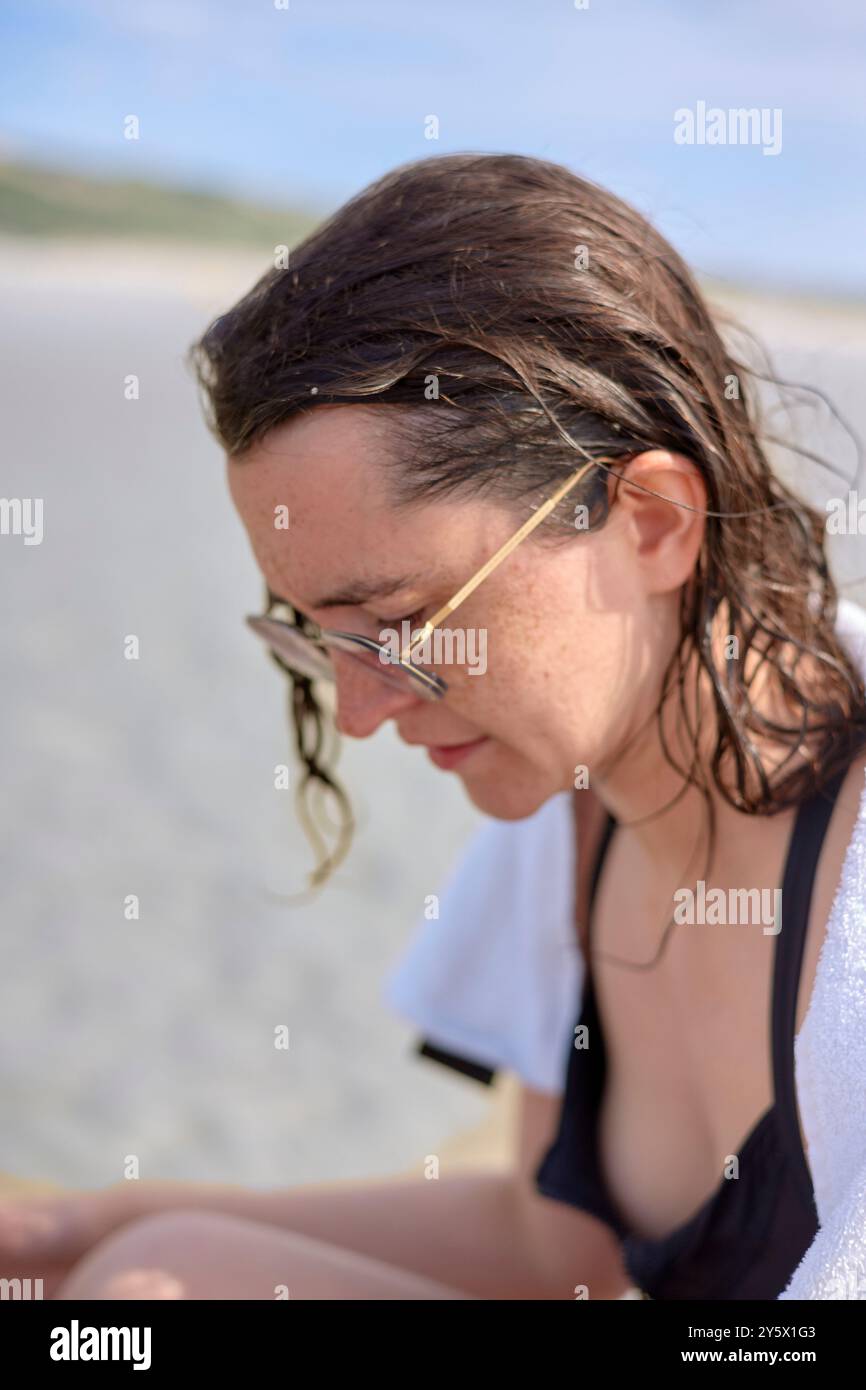 Junge Frau mit Brille am Strand, nachdenklich mit nassem Haar nach unten schauend. Stockfoto