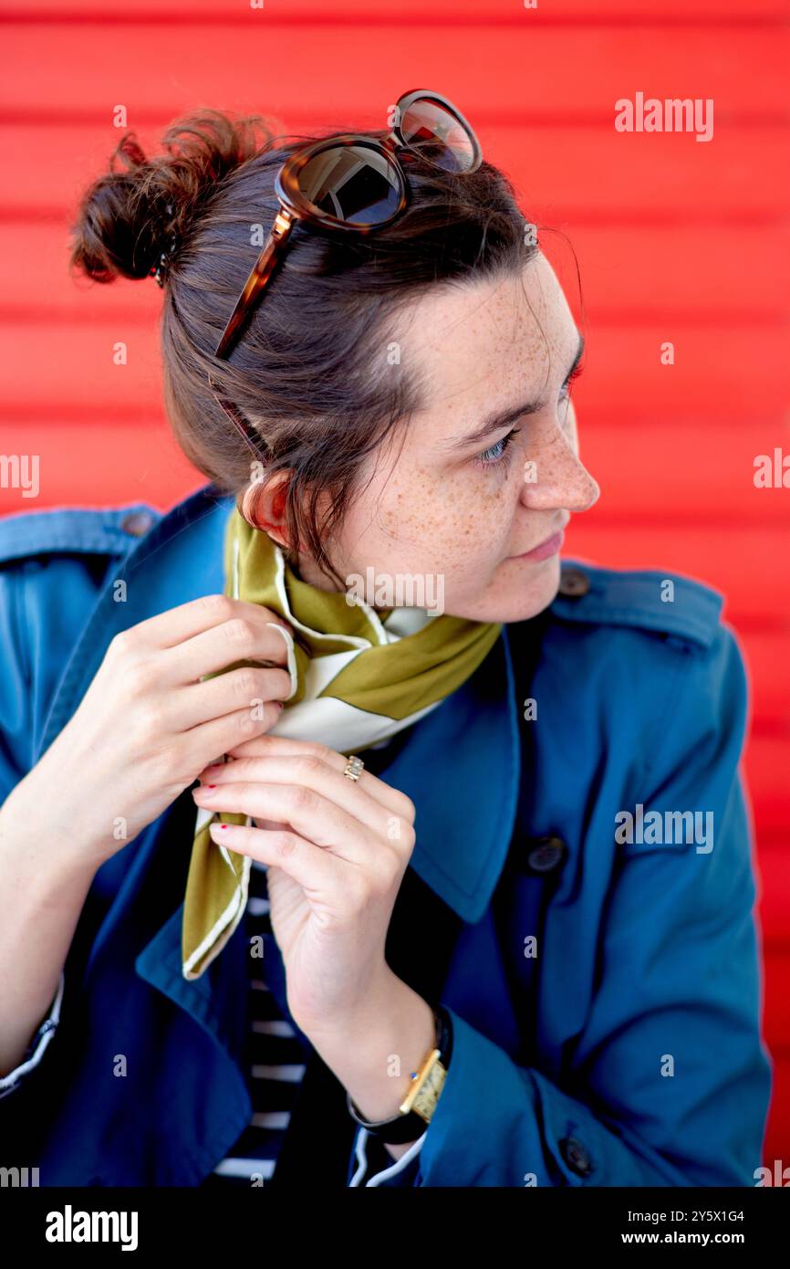 Frau in einer blauen Jacke, die ihren grünen Schal vor einem roten Hintergrund anpasst Stockfoto