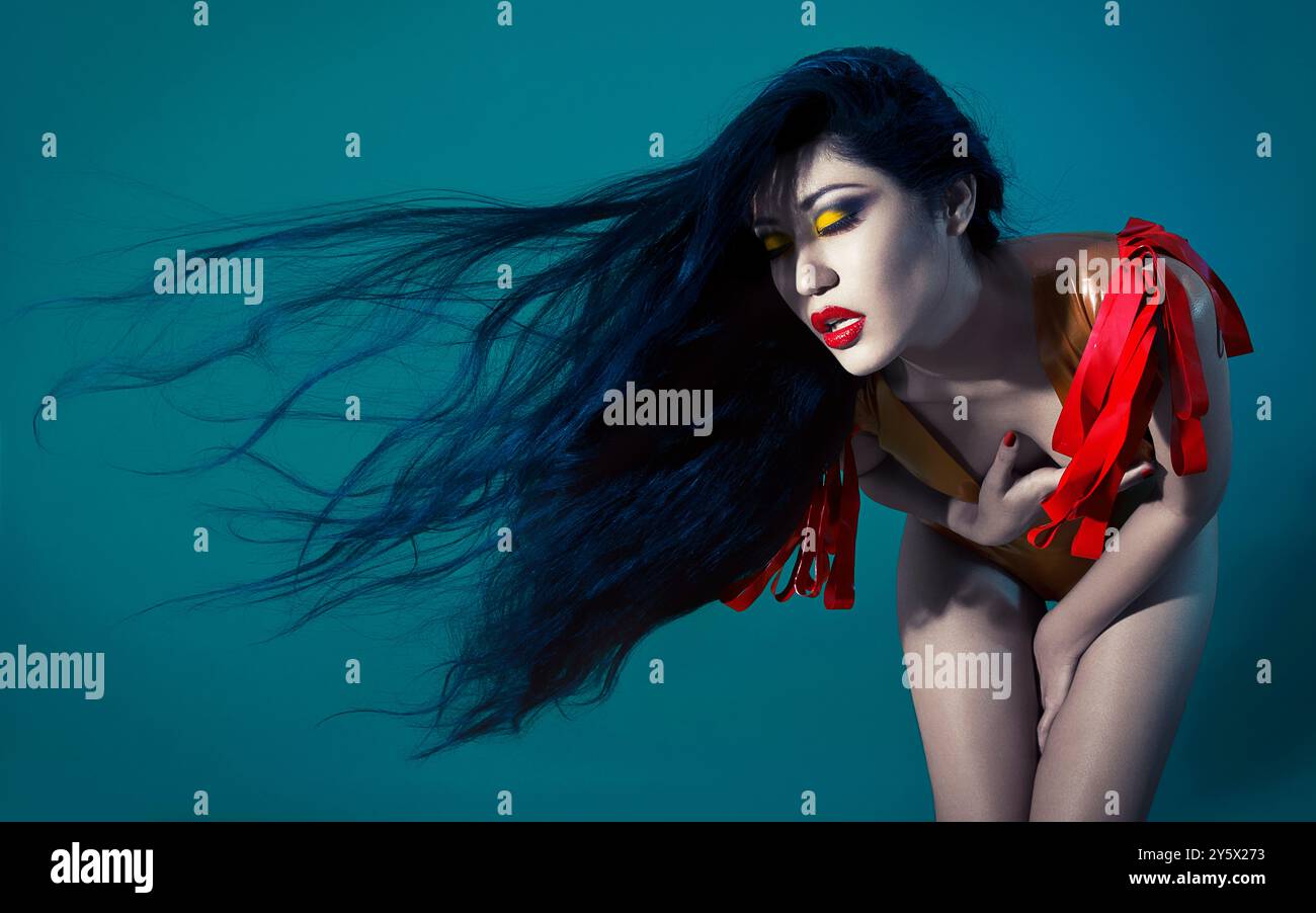 Frau mit langen, fließenden schwarzen Haaren und auffälligem gelbem Augen-Make-up mit rotem Harness auf blauem Hintergrund. Stockfoto