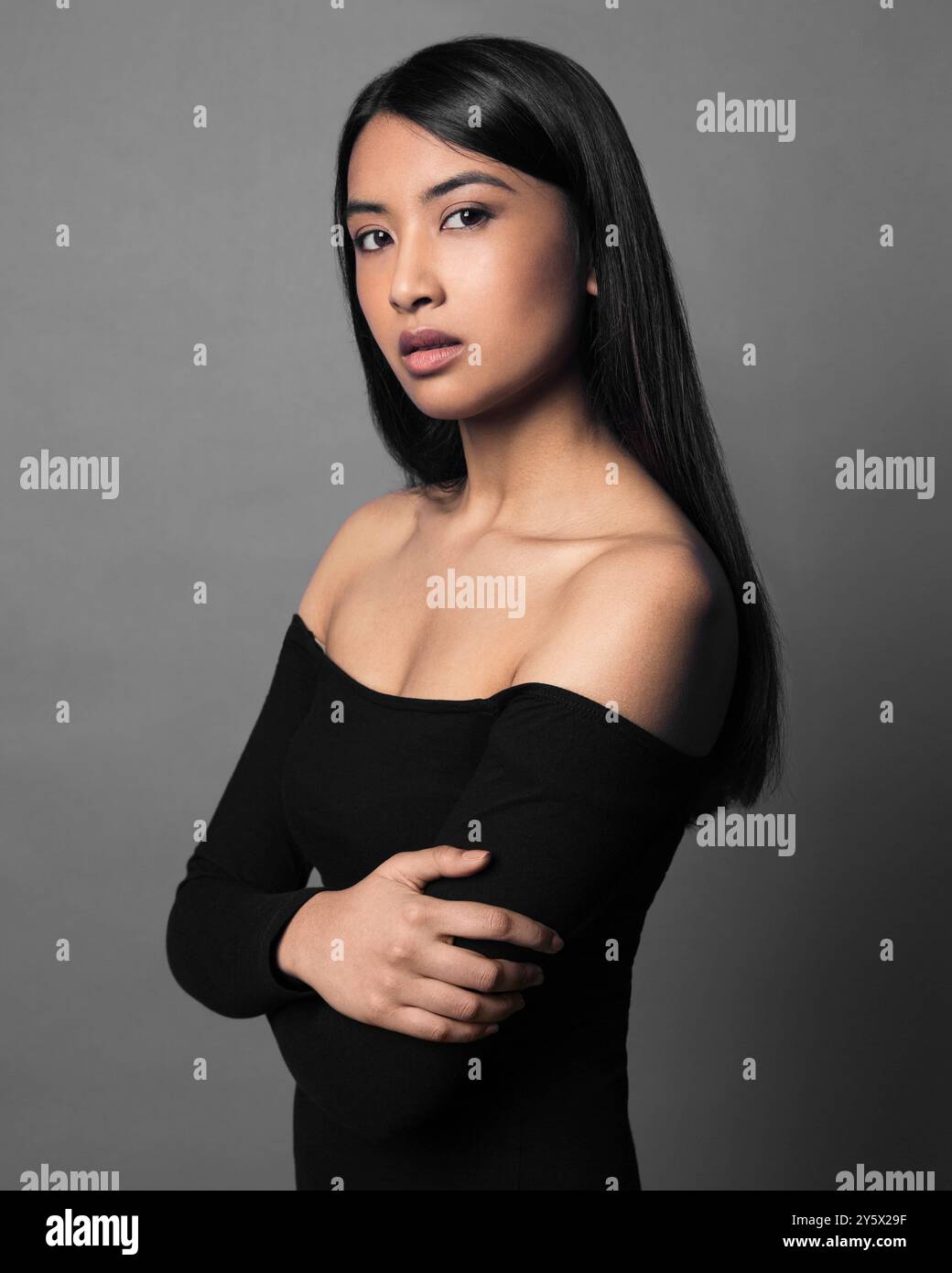 Porträt einer jungen Frau mit langen dunklen Haaren, die ein schwarzes Off-Shoulder-Top auf grauem Hintergrund trägt. Stockfoto