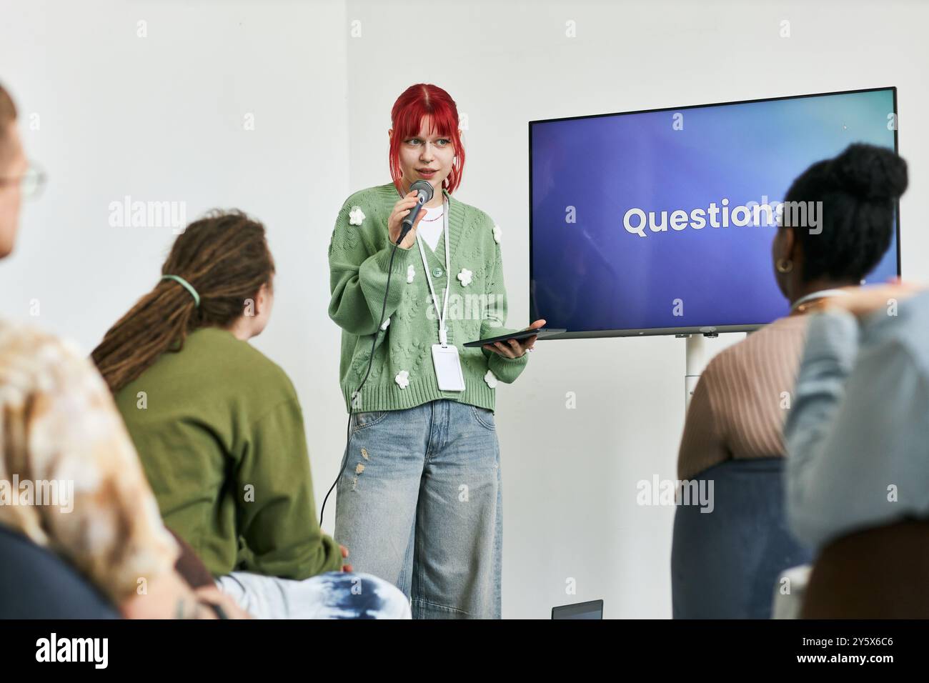 Redner mit rotem Haar, der das Publikum mit Mikrofon und Tablet anspricht, während er in der Nähe des Bildschirms steht und Fragen im Konferenzraum anzeigt, während verschiedene Teilnehmer an der Diskussion teilnehmen Stockfoto