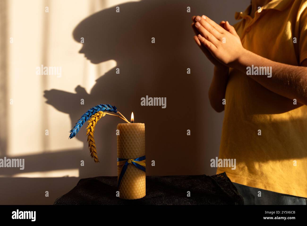 Brennende Kerze, zwei Weizenohren, gelb-blau gegen den Schatten des Kindes gemalt, mit gefalteten Händen im Gebet Stockfoto
