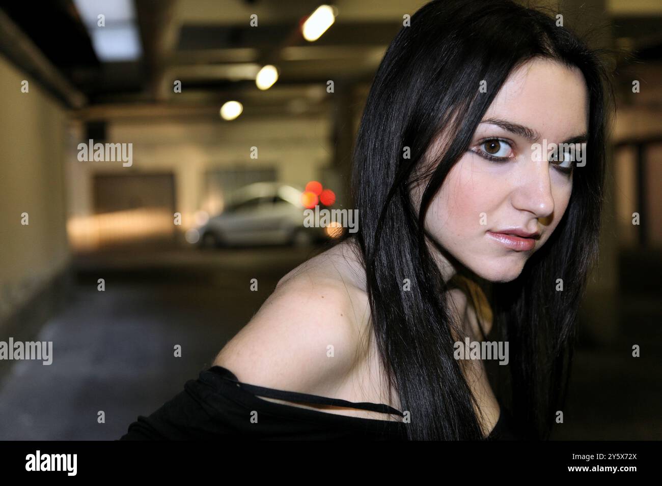 Frau mit dunklen Haaren, die ein schulterfreies Top trägt, posiert in einer schwach beleuchteten Garage mit einem Auto im Hintergrund, Mailand, Italien Stockfoto
