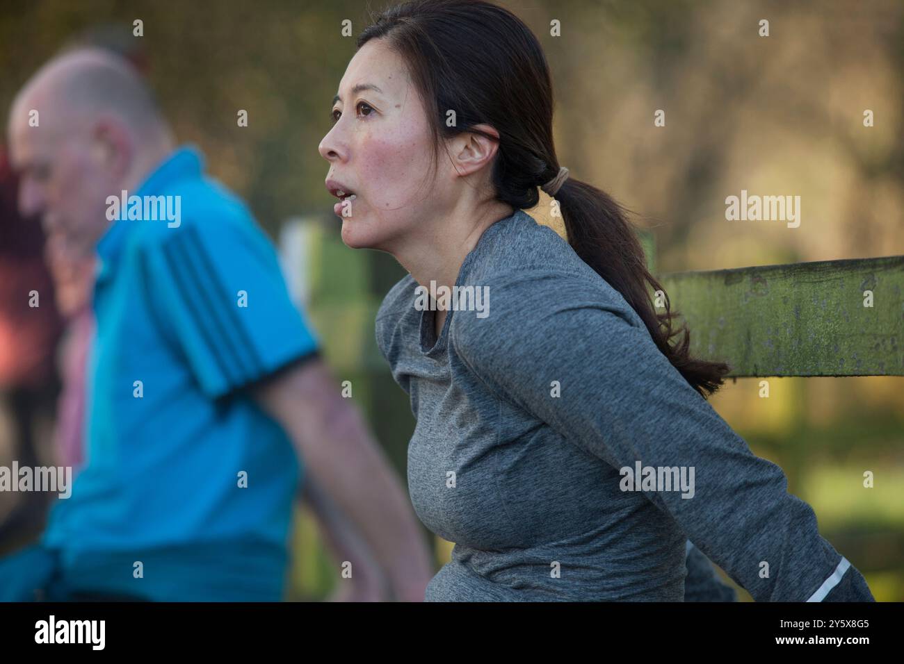 Eine Frau in einem grauen Oberteil liegt während eines Workouts auf einer Holzbank, während ein verschwommener Mann im Hintergrund joggt. Stockfoto