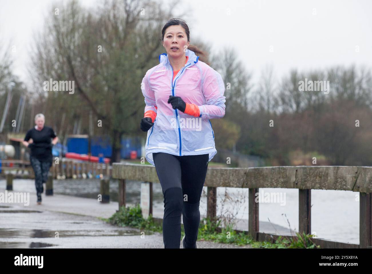 Frau, die in einem Park mit einem See joggt, und ein weiterer Läufer im Hintergrund an einem bewölkten Tag. Stockfoto